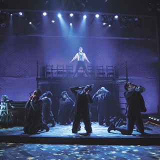 An a scene from TCU's presentation of the musical Spring Awakening, an ensemble of actors in period costume kneel while a male soloist sings under a blue spotlight on an elevated platform.