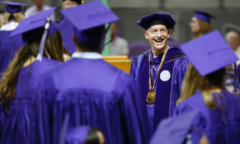 Chancellor Victor J. Boschini, Jr., smiling and in full academic regalia, greeting TCU graduates in purple caps and gowns