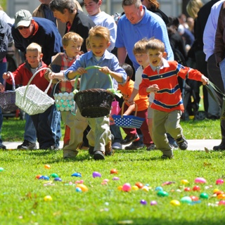 A group of young children holding Easter baskets run across a lawn covered with plastic eggs while their parents cheer behind them.
