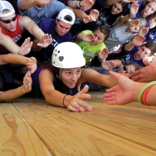 A young women wearing a protective rock-climbing helmet reaches up to the waiting hands of teammates as she scales a wooden outdoor obstacle course wall, surrounded by friends.