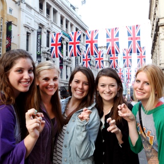 A group of five female TCU students studying abroad make the two-fingered Go Frogs hand sign while standing together on a street in London.