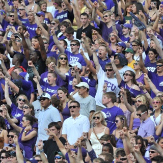 Dozens of cheering TCU fans raise their arm in a two-fingered "Go Frogs" hand sign at a crowded football game.