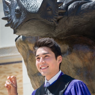 A male TCU student in Honors regalia makes the two-fingered "Go Frogs" hand sign in front of a bronze statue