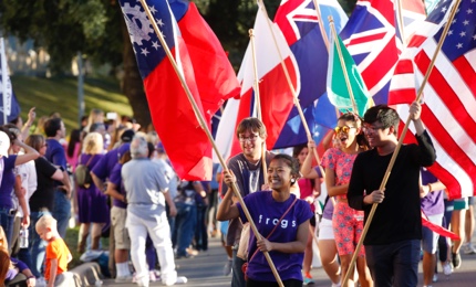 A group of laughing students carry flags of different nations in a TCU Homecoming parade, led by a girl wearing a shirt that says "Frogs"