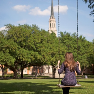 Back of female TCU student sitting in a metal swing with a cluster of trees and the TCU chapel in the background.