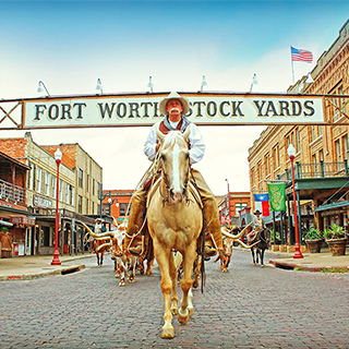 A cowboy in customary western attire rides a brown and white horse underneath a "Fort Worth Stock Yards" sign that spans across a street lined with old western buildings