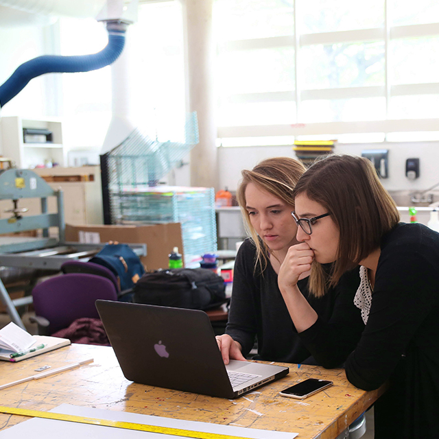 Two female TCU students view a laptop in the printmaking studio