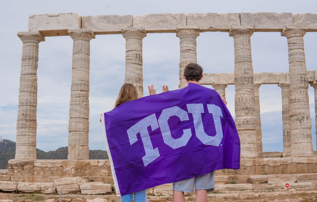 Two students, backs toward us, drape a TCU flag across their shoulders as they look at the pillars of a classical monument