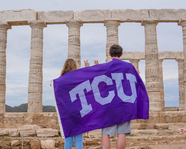 Two students, backs toward us, drape a TCU flag across their shoulders as they look at the pillars of a classical monument