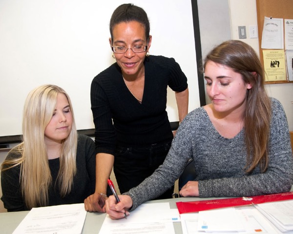 An economics professors looks on as one of two students writes out an idea on a piece of paper