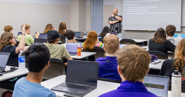A classroom full of students facing a projector screen. A professor at the front calling on a student with their hand raised.